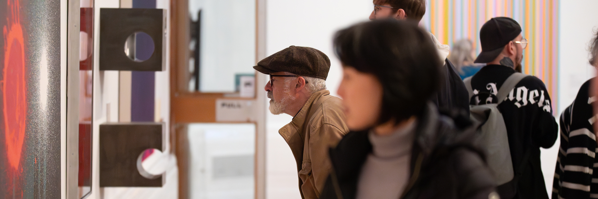 A photograph of two people looking at art in a gallery. One is stood upright, the other is leaning towards the wall.
