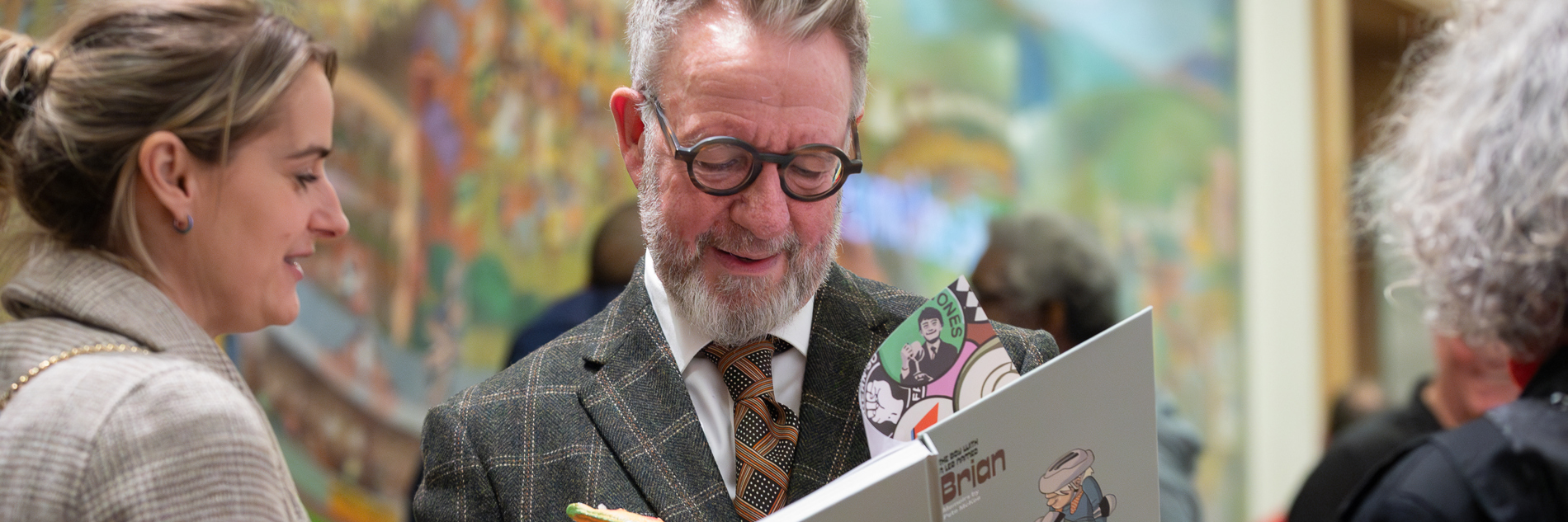 A photograph of an adult signing a book. An adult is standing next to them and other adults can be seen in the background.
